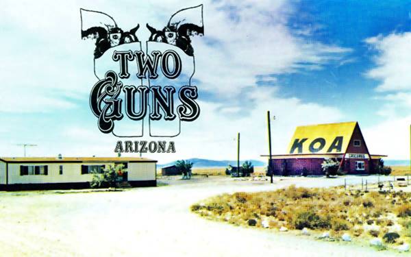 1970s poscard Two Guns KOA Steep pitched roof painted yellow, word KOA on it, desert, a low office to the left, shrubs