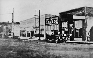 1910s black and white: garage buildings, dirt street, car