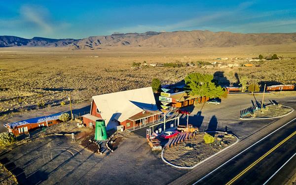 Old gas station left, former motel rooms right, green Tiki head left, trees. Hills in the distance, Route 66 lower right, aerial view