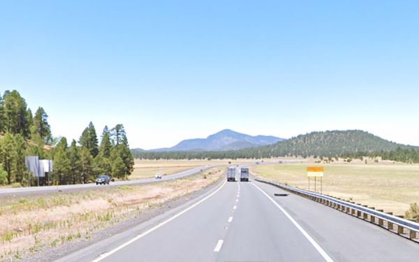 looking west along the freeway, trucks. Flat area ahead, forested hills and Bill Williams Mountain. Pine trees to the left