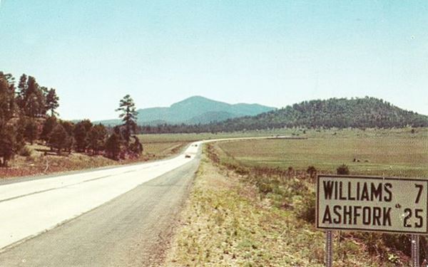 looking west along US 66, 1950s color photo, vehicles. Flat area ahead, forested hills and Bill Williams Mountain. Pine trees to the left