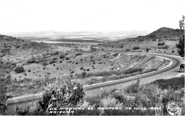 black and white postcard c.1940s: car on a long winding incline, hill to the right, slope to the left, butte on the horizon