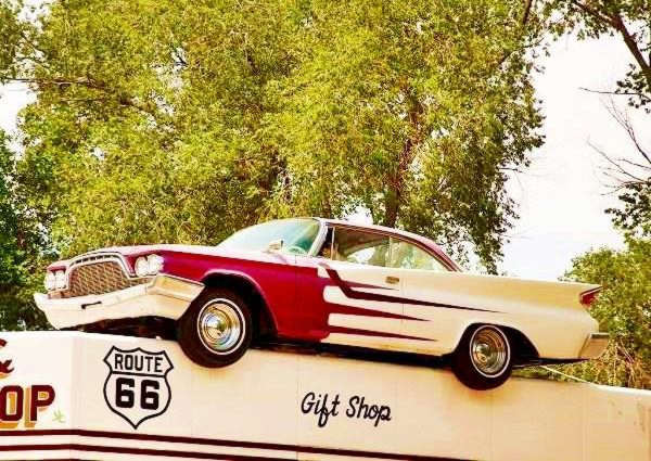 A red and white 1960s DeSoto with Elvis at the wheel, on the rooftop of a former gas station, treetops behind