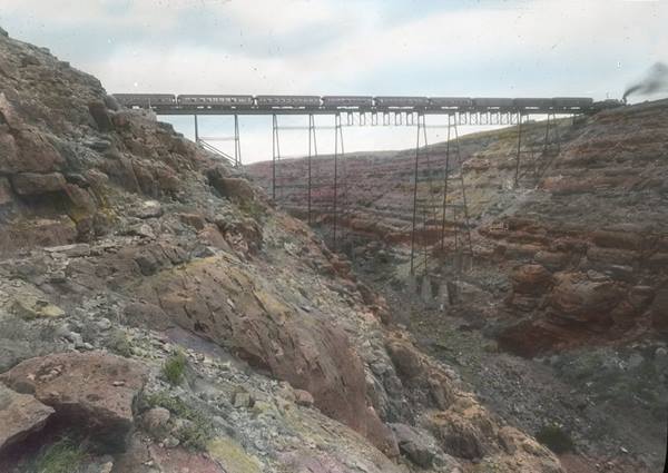 colorized late 1800s postcard of a steel bridge spanning a chasm, train with steam engine on the bridge