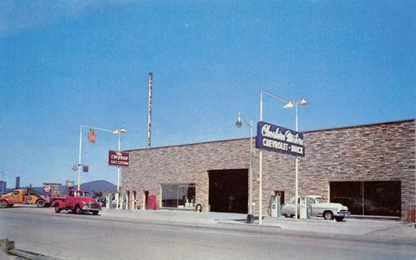 box shaped flagstone faced building, with service bays, gas pumps, tower truck and neon signs, 1950s postcard