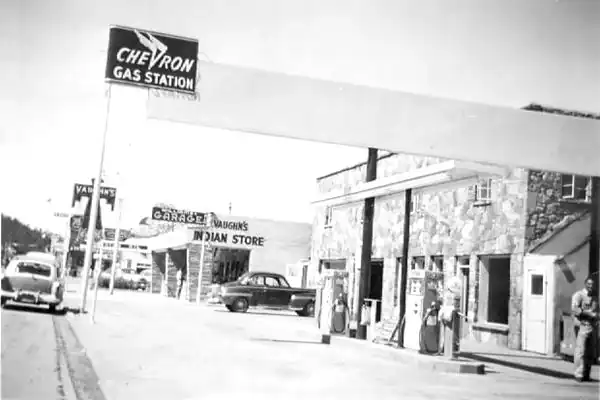 black & white 1950s photo: long white sign with the words:Chevron Gas Station on it, cars, gas pump, bldgs