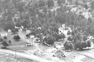 black & white aerial photo c.1930: US 66 on the bottom, gas station next to it, cabins among the pines on a hill