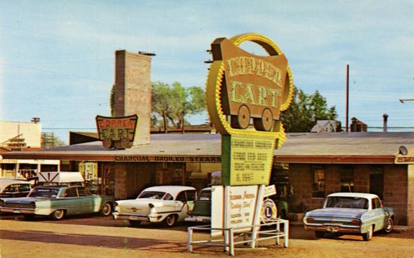 1960s cars in front of restaurant with neon sign shaped like a mining cart. Color postcard