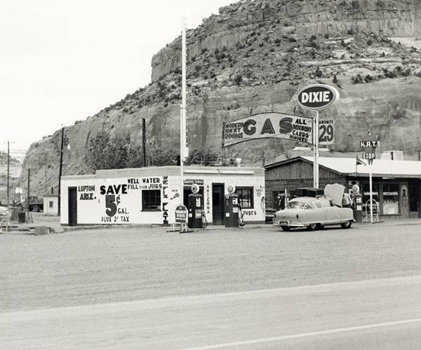 black and white photo Dixie El Paso block shaped gas station and pumps left, signage with gas price and long timber faced, gable roofed trading post and cars seen from old US66, in 1962