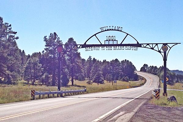 steel arch spans the highway "Williams Gateway to the Grand Canyon - 1881" written on it. Hill with pine trees in the background