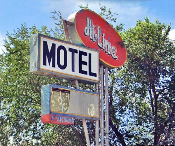 1950s neon sign: red oval with words " Hi-Line Motel" below a white box with black letters spelling MOTEL, lowest box has broken acrylic. Steel poles support it. Tree tops behind. 