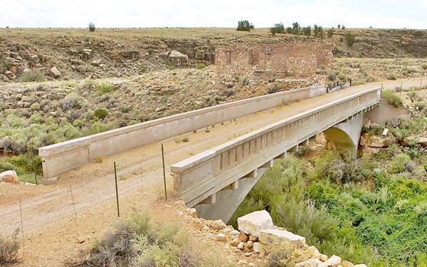 concrete bridge spanning canyon, dirt road across it, ruins in the background. Arid terrain with bushes