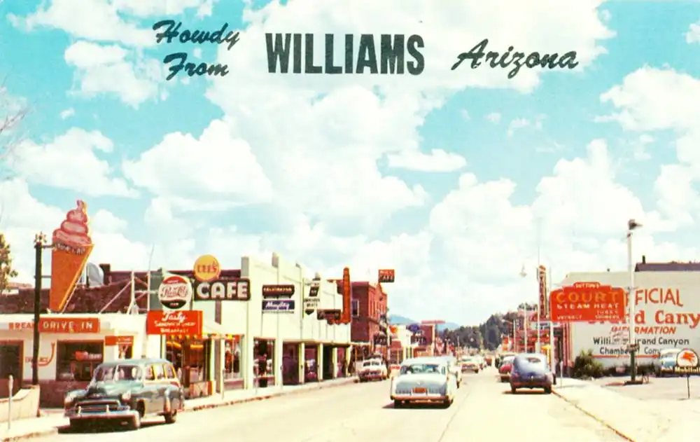1950s color postcard: "Howdy from Williams Arizona" looks east along Route 66, business district; cars, people, stores, neon signs