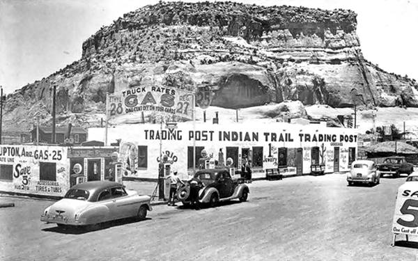 block shaped gas station and pumps left, signage with gas price and long stucco walled trading post and cars seen from old US66, a 1940s postcard