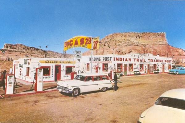 block shaped gas station and pumps left, signage with gas price and long stucco walled trading post and cars seen from old US66, 
a 1950s postcard
