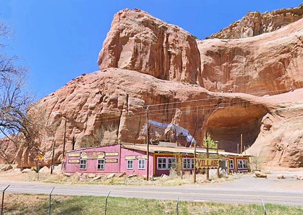 faded red gable roofed, vacant building with broken windows and trading post signs, by imposing sandstone cliff