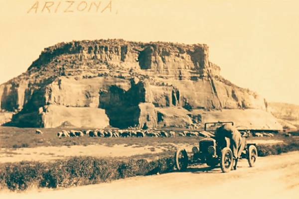 sepia image of man working on the motor of a 1920s car, sheep in a dry field next to a his car, sandstone cliffs beyond. Gravel surfaced US66 runs aceross the front. Car on the road