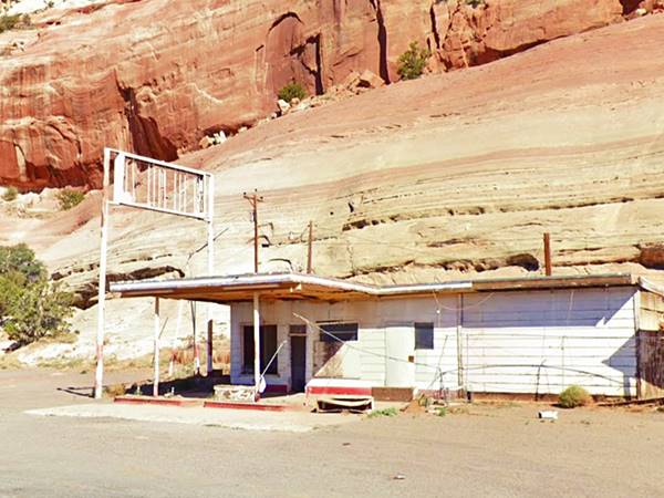 flat roof, flat canopy box shaped woodframe former gas station, broken sign above, red sandstone cliffs behind. gravel drive