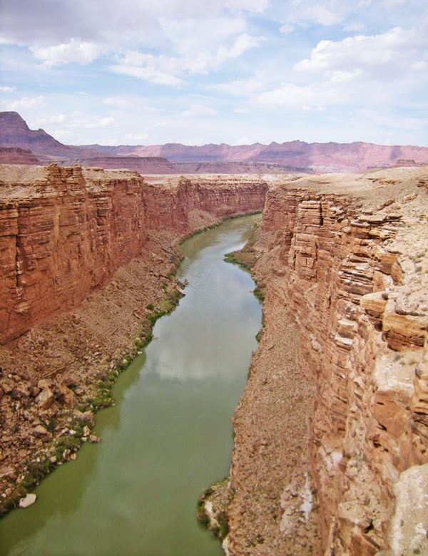 A narrow section of the Grand Canyon, tall vertical walls, green colored water below, the plateau and mountains in the distance