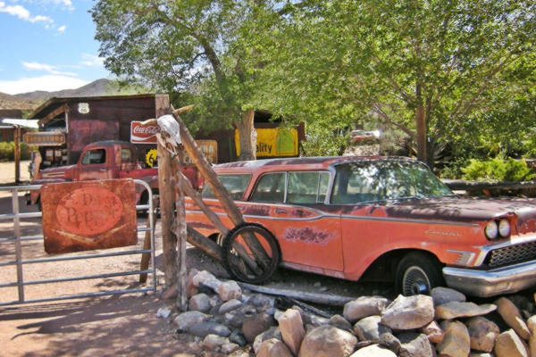 rusting sun faded 1960s station wagon and 1950s pickup truck under trees next to old gas station with vintage signs