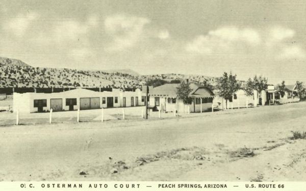 1930s black and white postcard, US 66 in the foreground, the auto court buildings with trees lined along the street. Hills in the background
