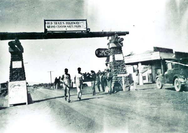two men run under a gateway with stone columns marking route as Old Trails and a reference to the Grand Canyon, General store to the right, cars, black and white 1928 photo