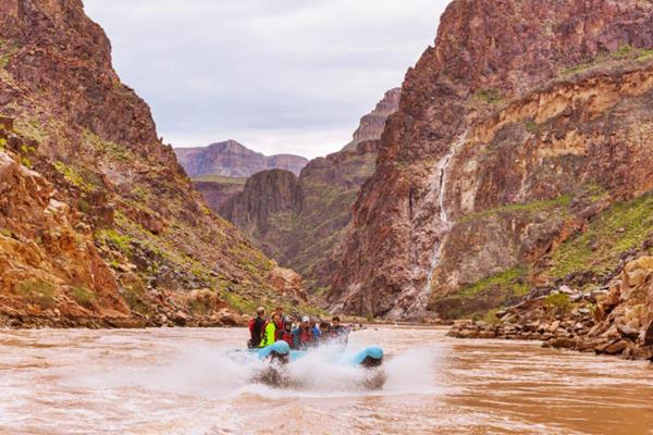 raft in the reddish-brown turbulent waters of the Colorado River, people with life jackets and helmets and steep rocky gorge enclosing the river