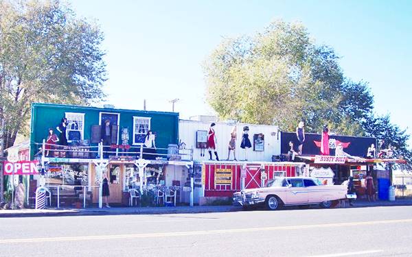 woodframe buildings, of different colors facing Route 66 with signs and mannequins decorating them and a pink Cadillac parked on the street