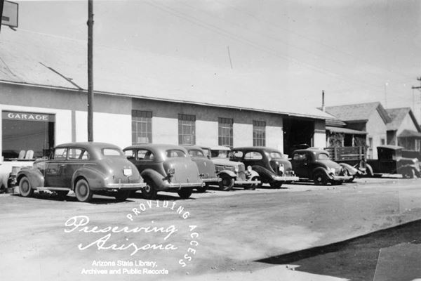 black and white postcard with 1940s cars lined up against stone wall of garage with multi-pane windows