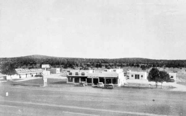 black and white postcard c.1950s: long, single floor gable roofed building to the left, words "Sleepy Hollow" written on the roof, central office and more units behind. Neon sign and 1950s cars. Route 66 runs in front