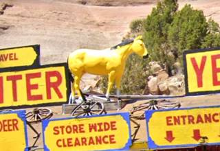 horse statue painted yellow on the roof of a trading post, signs, and cartwheel next to it, behind a juniper and red sandstone cliffs
