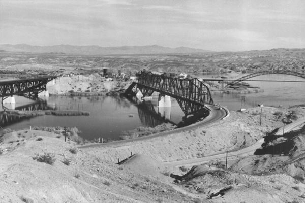 black and white photo looking east from California to Arizona, the Colorado river runs across the image and three bridges span it, two have roads coming from them. Arid hills