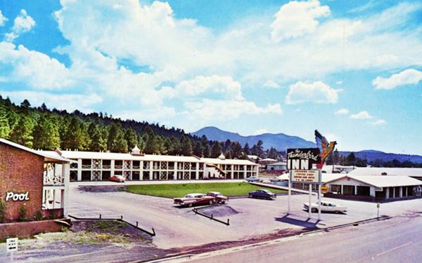 late 1950s color postcard of a two story motel with gable roof, wooded hill behind, Thunderbird shaped neon sign and cars in the parking lot