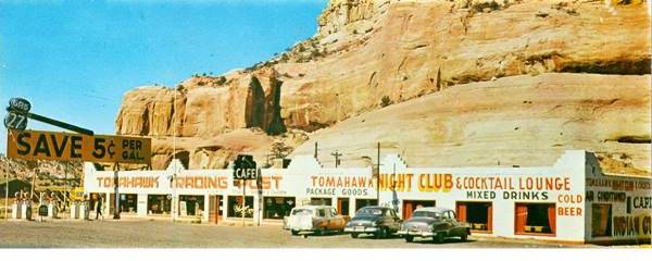 color postcard with gas pumps facing a long single story stucco building with a cafe, package goods, night club. Cliffs behind it and sign promising gas 5 cents per gal. savings