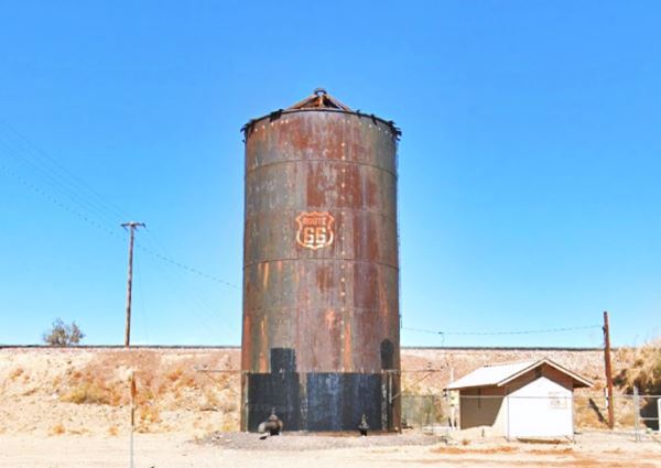 rusty tank with a white US66 shield, a railroad embankment behind it, seen from I-40, arid surroundings, blue sky, bushes