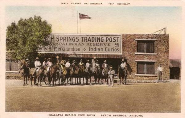 A colorized 1936 postcard showing the Trading Post with cowboys on horseback posing in front of it