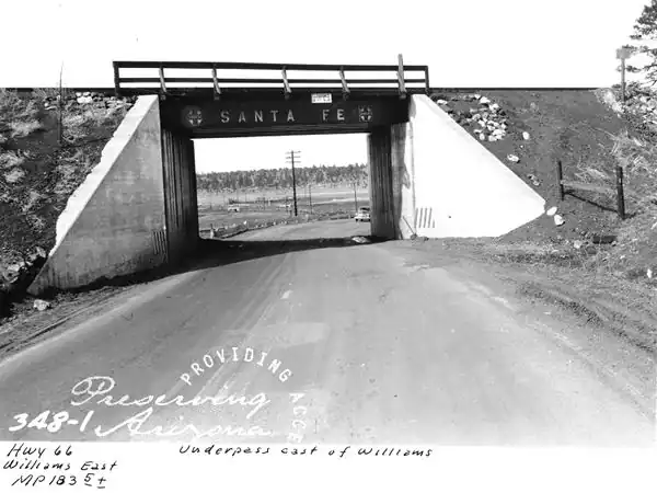 black and white, 1940s car on US 66 reaching railroad underpass, hill beyond