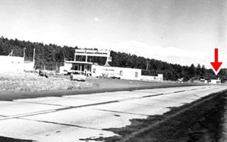 black and white photo a gas station, cars, Route 66 in the foreground and a red arrow marking a motel in the distance. Forested hills in the background