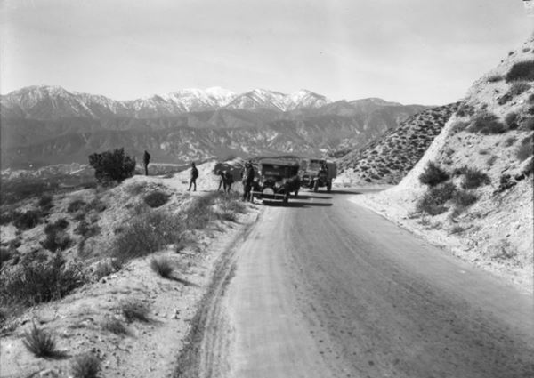 National Old Trails hwy in Cajon Pass, 1920 black and white, 1920, men and cars on a narrow road, arid scenery at a lookout, snowcapped mountains in the distance