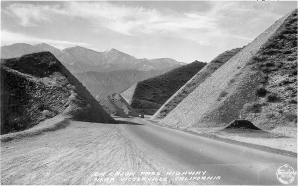 US66 in Cajon Pass, 1931 black and white, 1931, car driving a highway flanked by deep cuts in mountain range, snowcapped mountains in the distance