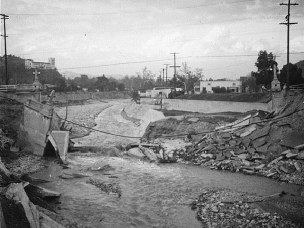 washed out 43 Ave. bridge, 1938 flood, Los Angeles CA black and white photo, 1938, ruins of bridge across a river with boulders and water. Hill in the distance with a building on it