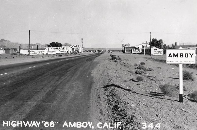 black and white 1940s view eastwards along US66, right: roadsign says AMBOY, cabins and cafe, gas station, middle: highway, left: signs of a Texaco, garage and building