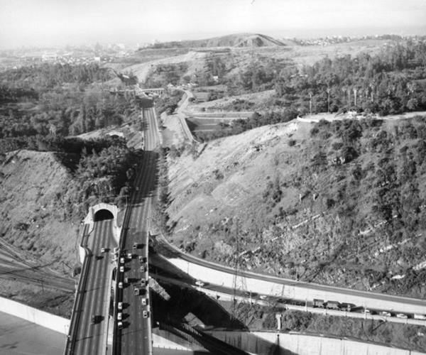 1958 Arroyo Seco Parkway at Elysian Park Los Angeles CA Route 66 aerial view black and white photo, 1958 mountains with vegetation, divided highway enters it on the left using tunnels, on the right with a cut. In the distance smog and Los Angeles downtown, in the foreground traffic by a river that runs left to right