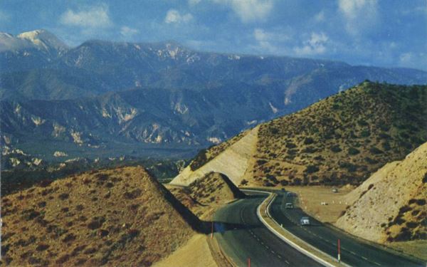 4-lane US66 in Cajon Pass, 1950s color, 1950s. Traffic along a 4 lane highway with median dividing east and westbound traffic. Cut in mountainside, snowcapped mountains in the distance