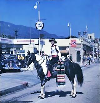 Union 76 c.1940s, Monrovia CA man on horseback as a cowboy in a parade, behind 1940s cars and a Union 76 gas station. Color, buildings, street lights, mountain behind