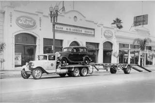 black and white, 1936. Trailer with car on it parked by a building with 5 large glass windows facing the street two FORD logos over the windows left and right AHRENS written over the central ones
