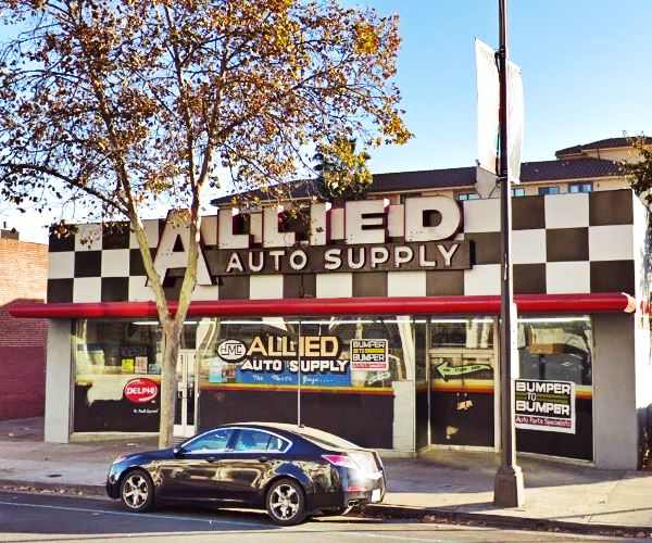 Car parts store, with sign spelling ALLIED AUTO SUPPLY on a black and white checkered panel, car parked in front, trees, street