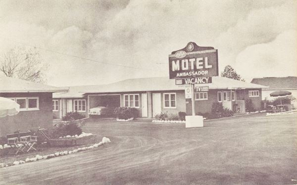 black and white 1950s. postcard: motel sign right, by a hip roof unit, with roofed garage included, clouds beyond, route 66 runs in front, parasol to the left
