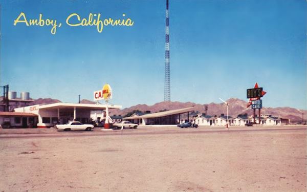 left to right, gas station,Shell, cafe with slanting roof, motel units (cabins) and neon sign, cars and US 66 in front, hills and radio antena behind 1960 color postcard