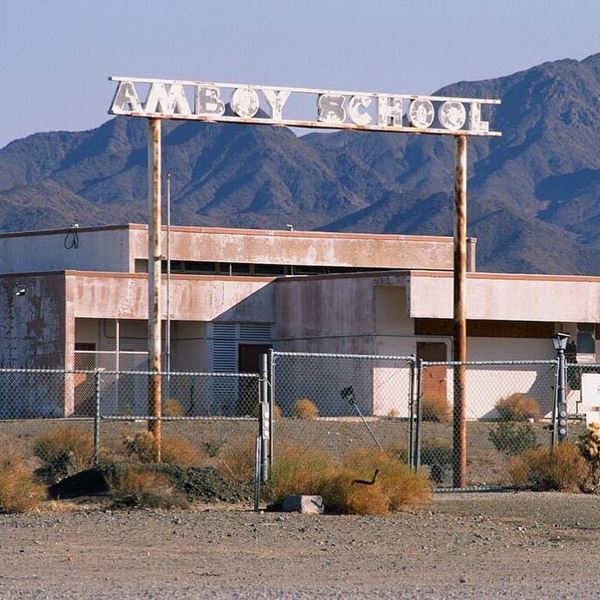 Amboy’s closed school US 66 gateway steel, faded peeling reads AMBOY SCHOOL, building behind, hills beyond, gate and fence padlocked